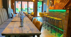 Image of a restaurant with wooden chairs and tables. Menus have been placed on top of the tables. Bar with bar stools and neon green light